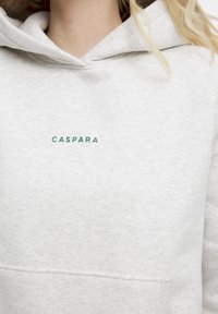 Ljusgrå hoodie i mjukt, texturerat tyg. Har en grön logotyp "CASPARA" centrerad på bröstet och en framåtriktad känguruficka.