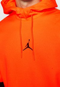 Felpa con cappuccio in tessuto arancione brillante con una superficie liscia. Presenta il logo nero di Jordan sul petto e cordini abbinati.
