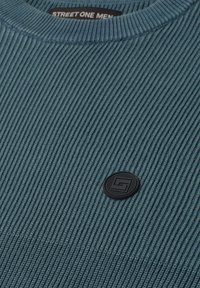 Tissu tricoté côtelé bleu avec un col rond et un petit patch logo circulaire noir sur la poitrine.
