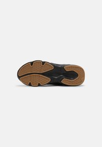Semelle de chaussure noire avec des crampons en caoutchouc marron présentant des motifs géométriques et des rainures profondes pour l'adhérence, montrée sur fond blanc.