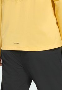 Geel lichtgewicht sporttopje met een gladde textuur, met een subtiel logodetail bij de zoom, gecombineerd met donkergrijze broek.