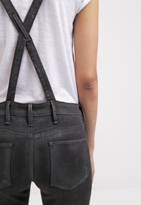 Black denim overalls with cross-back straps, fitted design, and slightly distressed texture. Features two back pockets and belt loops.