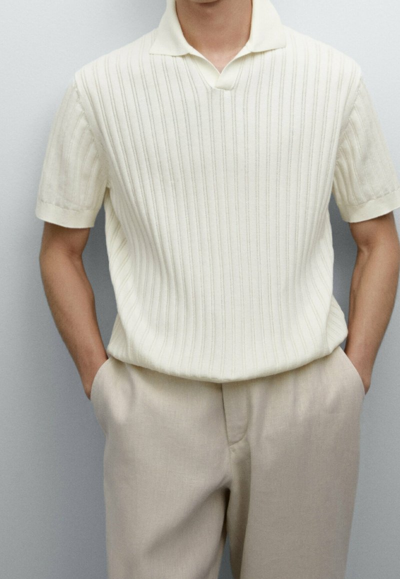 Homme portant un polo en tricot côtelé blanc à manches courtes rentré dans un pantalon en lin beige, les mains dans les poches, sur un fond uni.