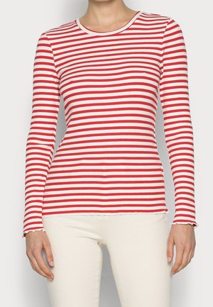 Kobieta ubrana w czerwono-białą koszulę z długim rękawem w poziome paski z wykończeniem w kształcie karbowanej sałaty oraz kremowe spodnie, stojąca na białym tle.