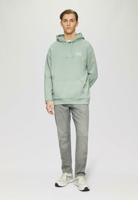 Felpa verde chiaro con tasca a marsupio, cappuccio regolabile con cordino e logo discreto. Abbinata a jeans grigi e sneakers bianche.