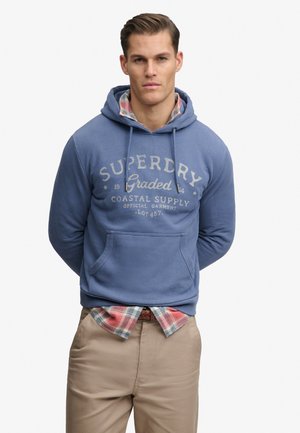 Mężczyzna w niebieskiej bluzie Superdry z kapturem na czerwonej koszuli w kratę, beżowe spodnie, stoi ze złożonymi za plecami rękami na białym tle.