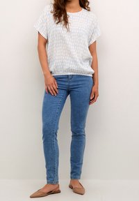 Kvinde iført lyseblå mønstret kortærmet top, blå stramme jeans og beige spidse ballerinaer, der står op ad en ensfarvet væg.