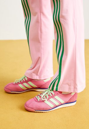Piedi con sneakers Adidas rosa e verde chiaro con lacci verde chiaro, abbinati a pantaloni rosa con tre strisce verdi sui lati.