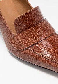 Mocasín de cuero marrón con textura de cocodrilo, que cuenta con una cinta ancha en el medio del pie y un diseño de punta afilada. Forro interior suave.