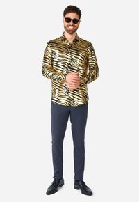Chemise à motifs zèbre en or et noir brillants avec un col, assortie à un pantalon texturé foncé et des chaussures de ville noires ; lunettes de soleil portées.