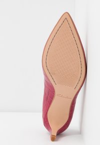 Chaussure rouge à bout pointu avec une tige texturée effet crocodile, une semelle marron clair lisse contrastante et un détail logo embossé subtil sous la semelle.