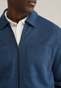 Zilā zivju kaulu raksta jaka ar rāvējslēdzēju, apkakli un divām priekšējām kabatām, vilkta pāri teksturētai baltai kreklai.