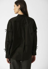Black button-up shirt featuring multiple fabric bows on the back and sleeves. Made of lightweight material with a relaxed fit.