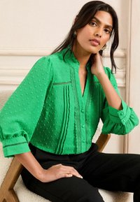 Blusa verde con pois texturizzati, maniche arruffate e dettagli in pizzo; presenta una chiusura a bottoni sul davanti e un colletto alla cinese.