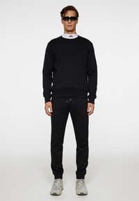 Set di felpa nera e pantaloni. Tessuto morbido, vestibilità rilassata, polsini a coste. Indossato con sneaker grigie e occhiali da sole scuri, maglietta bianca semplice visibile.