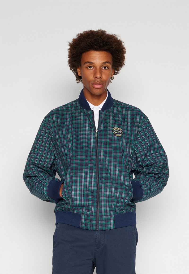 Veste bomber à carreaux en vert et bleu marine, avec une fermeture éclair à l'avant, des poignets côtelés et un patch logo. Le matériau semble léger et texturé.