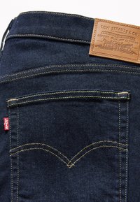 Jean en denim foncé avec coutures dorées, doté d'un patch en cuir portant l'inscription "Levi Strauss & Co." et d'une petite étiquette rouge sur la poche arrière.