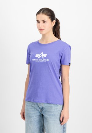 Jeune femme aux cheveux bruns en queue de cheval, portant un t-shirt violet Alpha Industries et un jean bleu clair, légèrement tournée vers la droite.