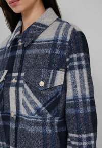 Vrouw in een marineblauw, grijs en lichtblauw geruit wollen jas met knopen en klepzak op een neutrale achtergrond.