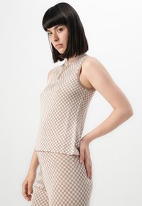 Žena s ravnom tamnom kosom odjevena u bezrukavnu bež i bijelu kariranu bluzu te odgovarajuće hlače, pozira ispred jednostavne bijele pozadine.