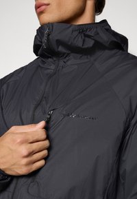 Moška roka dviga zadrgo na prsnem delu črne jakne Peak Performance z visokim ovratnikom.