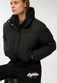 Czarna puchowa kurtka z wysokim kołnierzem i bocznymi kieszeniami, z pikowanym wzorem i zapięciem na zamek. Nosi się ją na czarnej bluzie z kapturem i dresowych spodniach.