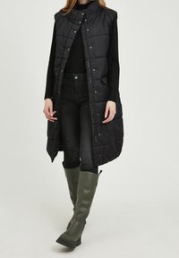 Černá nadýchaná bezrukávová vesta s vysokým límcem a patentkami, nošená přes černou košili s dlouhým rukávem a tmavé džíny, doplněná zelenými botami.