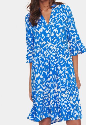 Femme portant une robe à motifs bleus et blancs, longueur genou, manches cloche trois-quarts et col en V, debout devant un fond uni.