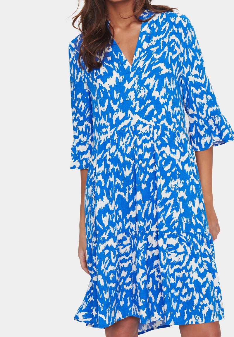 Femme portant une robe à motifs bleus et blancs, longueur genou, manches cloche trois-quarts et col en V, debout devant un fond uni.