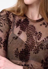 Close-up van een persoon die een transparant bruin shirt draagt met donkere, fluwelen bloemenpatronen en zichtbare huid op de borst en armen.