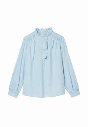 Donna che indossa una camicetta azzurro chiaro con colletto arricciato e pantaloncini beige a vita alta con tasche con bottoni, in piedi davanti a uno sfondo semplice.