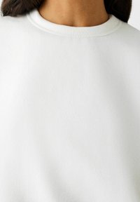 Felpa bianca in tessuto strutturato, con scollatura rotonda e bordi a coste. La superficie è liscia e uniforme, senza ulteriori motivi o decorazioni.