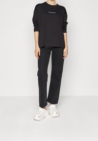 Camiseta negra de manga larga con un logo blanco, combinada con unos vaqueros oscuros de corte holgado y zuecos blancos con orificios de ventilación y suelas gruesas.