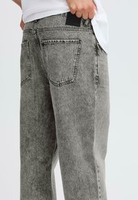 Persona que viste jeans de denim gris desgastados con bolsillos traseros, levantando ligeramente una camisa blanca para revelar una etiqueta negra sobre el bolsillo.