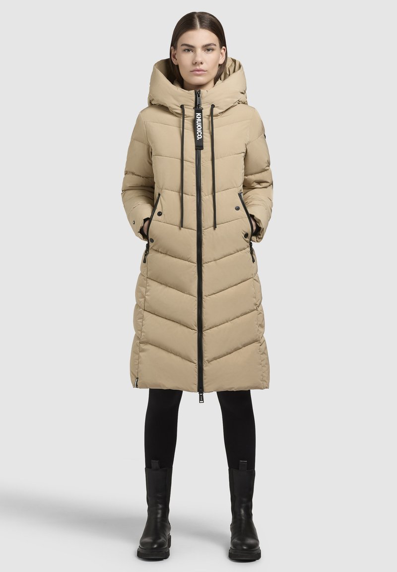 Beige pitkä untuva takki, jossa on chevron-kuvio, säädettävä huppu, musta vetoketju ja sivutaskut. Päällä mustat leggingsit ja saappaat.
