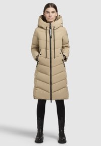 Manteau doudoune beige avec capuche, présentant un design en chevrons et des zips noirs. Porté sur un legging noir et des bottines.