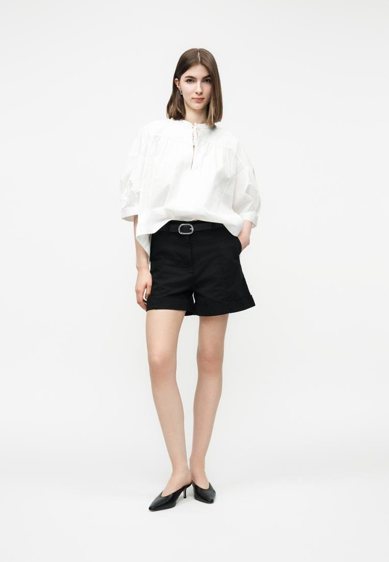 Jeune femme debout portant une blouse blanche oversize, un short noir ceinturé et des chaussures à talons noires à bride arrière sur un fond blanc uni.