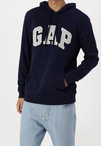 Sudadera azul marino de algodón, con un bolsillo frontal, capucha con cordón y el logo "GAP" en gris claro en el pecho.