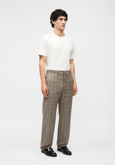 Άνδρας με σγουρά μαλλιά στέκεται φορώντας μια απλή λευκή κοντομάνικη μπλούζα, καφέ καρό παντελόνια με κορδόνι και μαύρα δερμάτινα παπούτσια.