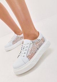 Sneakers bianchi con un pannello laterale testurizzato e decorato in beige e accenti argentati. Presentano lacci arcuati e una suola bianca spessa e testurizzata.