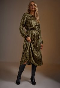 Vestido de estampado de leopardo en verde oliva, con mangas largas, cintura ceñida y dobladillo a media longitud. Presenta una textura sedosa combinada con botas de cuero negro hasta la rodilla.