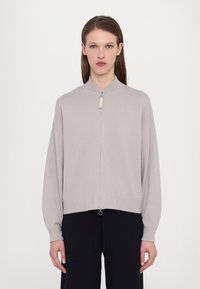 Ljusgrå zip-up tröja med mandarinhuvud, sänkta axlar och ribbade manschetter, med en silverfärgad dragkedja och ett något strukturerat tyg.
