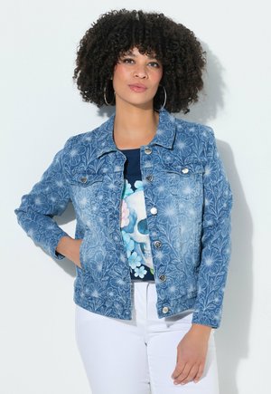 Kobieta z kręconymi włosami, nosząca kolczyki koła, niebieską kurtkę dżinsową w kwiatowy wzór nałożoną na koszulkę z nadrukiem czaszki oraz białe spodnie, pozująca z jedną ręką w kieszeni.