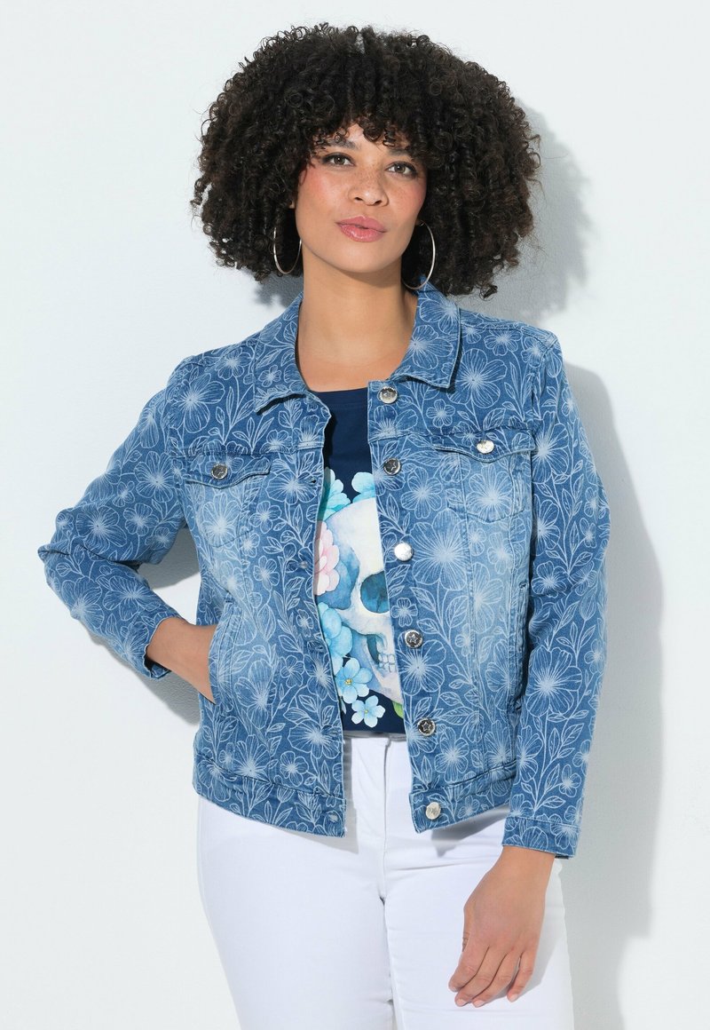 Frau mit lockigem Haar, die Creolen trägt, eine blaue Jeansjacke mit Blumenmuster über einem schädelbedruckten Shirt und weiße Hosen, posiert mit einer Hand in der Tasche.