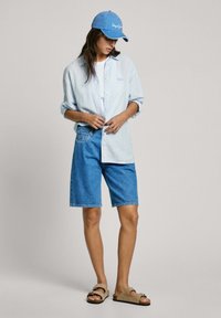Heledad sinine triibuline särk, valge t-särk, sinised denim-lühikesed püksid, beežid sandaalid, sinine müts logo jahedas vabas stiilis riietus.