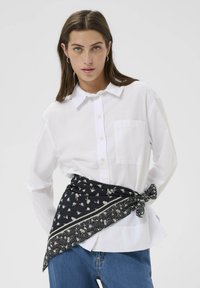 Camisa blanca de botones con un bolsillo, combinada con una bufanda de patrón floral negro atada en la cintura. Ajuste casual, textura suave, mangas de tres cuartos.
