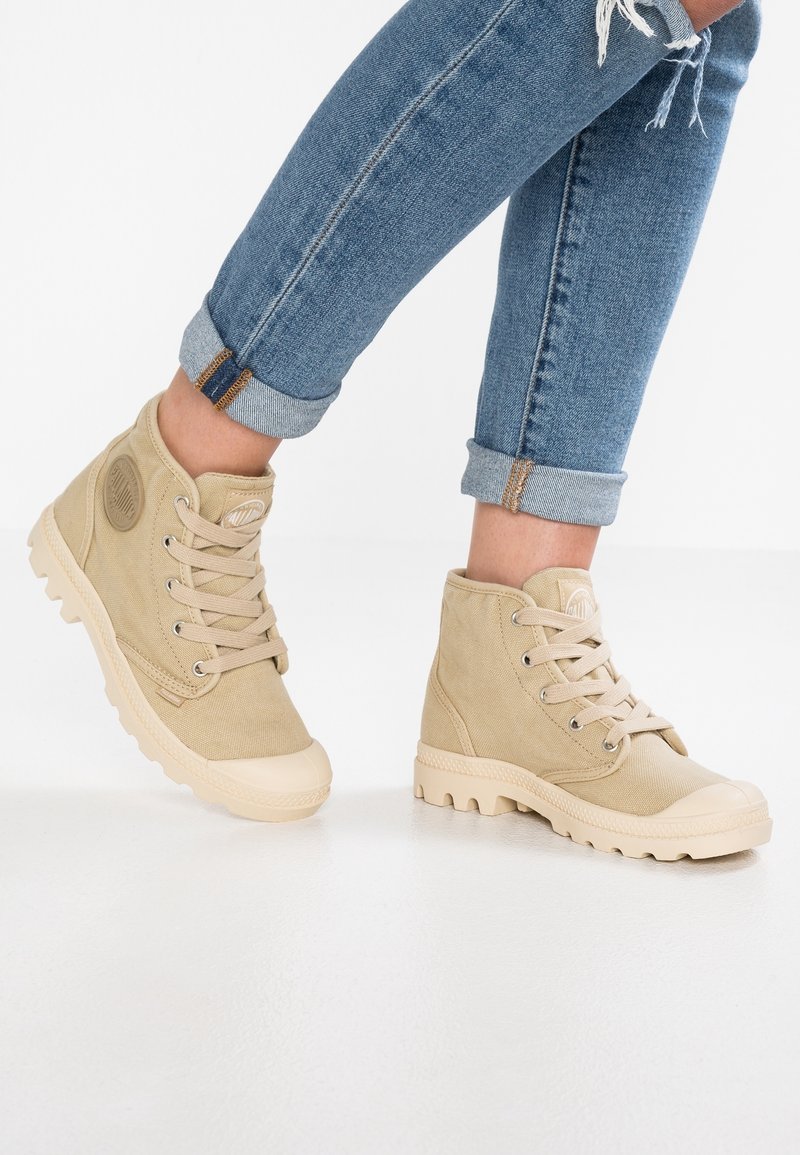 Bežové plátěné tenisky s vysokým střihem, gumovou podrážkou, kulatou špičkou a šněrováním vpředu. Obsahují logo a strukturované textilní akcenty.