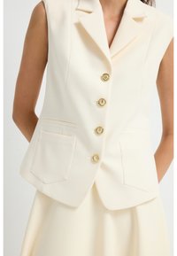 Elfenbensfarvet ærmeløs blazer med opslået krave, fem guldfarvede knapper og to forlommer. Glat stof med skræddersyet pasform.