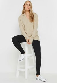 Maglione beige a trama cable-knit con scollatura a V e collare, abbinato a leggings neri e sneakers bianche. Texture liscia, vestibilità comoda, design minimal.