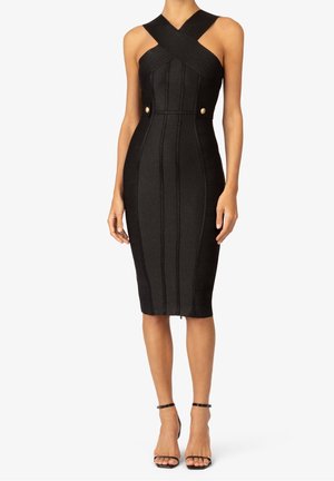 Femme portant une robe noire ajustée longueur genou avec des bretelles croisées et des boutons dorés à la taille, associée à des talons noirs à bride à la cheville.